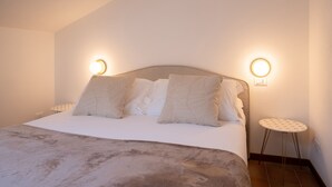 Un lit bien fait, avec une tête de lit, deux tables de chevet dotées de lampes rondes, et une petite table ronde recouverte d’une nappe à motifs.