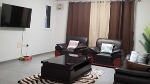 A living room with a flat-screen TV, two leather sofas, a coffee table, and a large window with curtains. - Escape to Eden heights,a 2 bedroom Apt with Gym access near the West Hills Mall. (Weija, Accra)