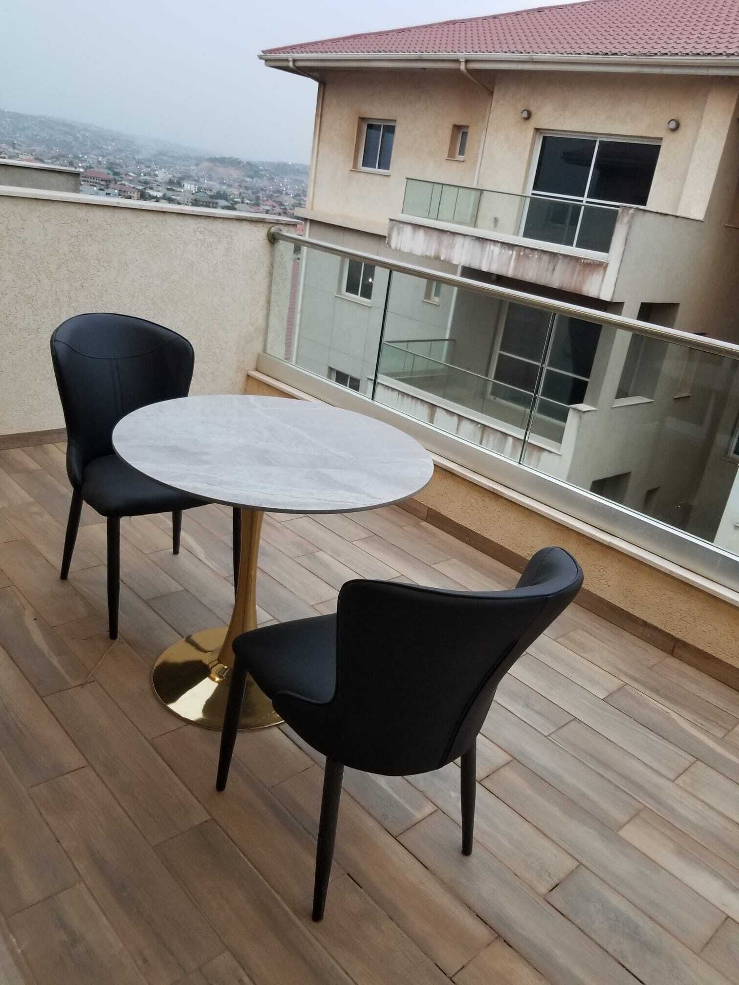 Ein Balkon mit rundem Tisch, zwei Stühlen, einem Glasgeländer und einem Gebäude im Hintergrund.