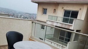 A balcony with a round table and two chairs, a glass railing, and a building in the background. - Escape to Eden heights,a 2 bedroom Apt with Gym access near the West Hills Mall. (Weija, Accra)