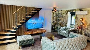 A modern living room with a large flat-screen TV, a staircase with wooden steps, a tufted sofa, and a decorative wall mural. - La Bastide du Causse - Domaine Gouloumès (Ginouillac)