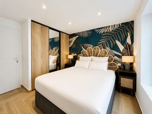 Un dormitorio moderno con una cama grande, un armario de madera, una pared con estampado de hojas tropicales y dos mesitas de noche con lámparas.