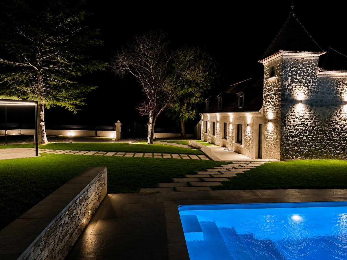 A nighttime view of a property with a swimming pool, stone walls, and a pathway leading to a building with multiple windows.