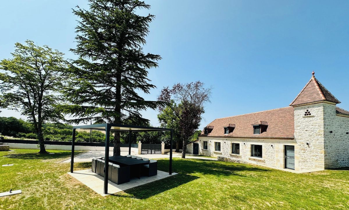 Ein Steinhaus mit Satteldach und einem Turm, umgeben von einem gepflegten Rasen und Bäumen.