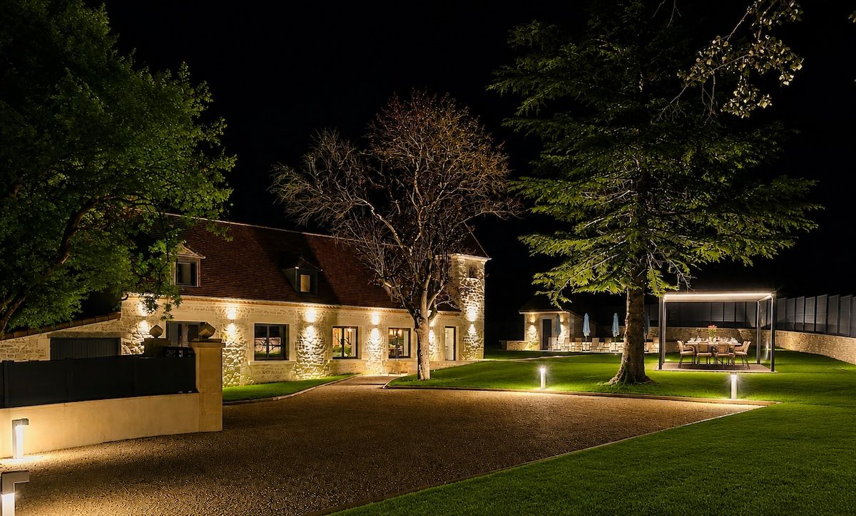 A stone house with a well-lit driveway, a covered outdoor dining area, and a lawn.