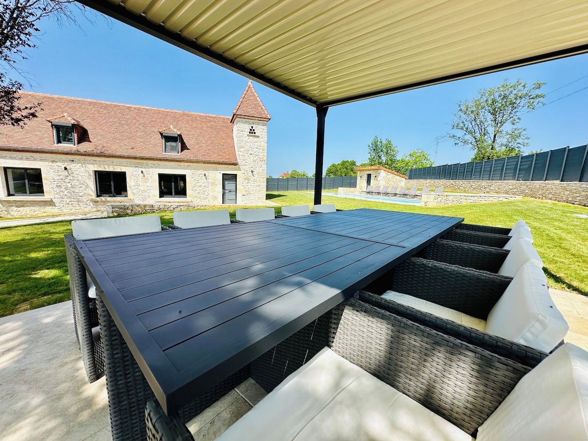 En udendørs terrasse med et langt bord og fletstole, et overdækket område og en bygning i sten i baggrunden.