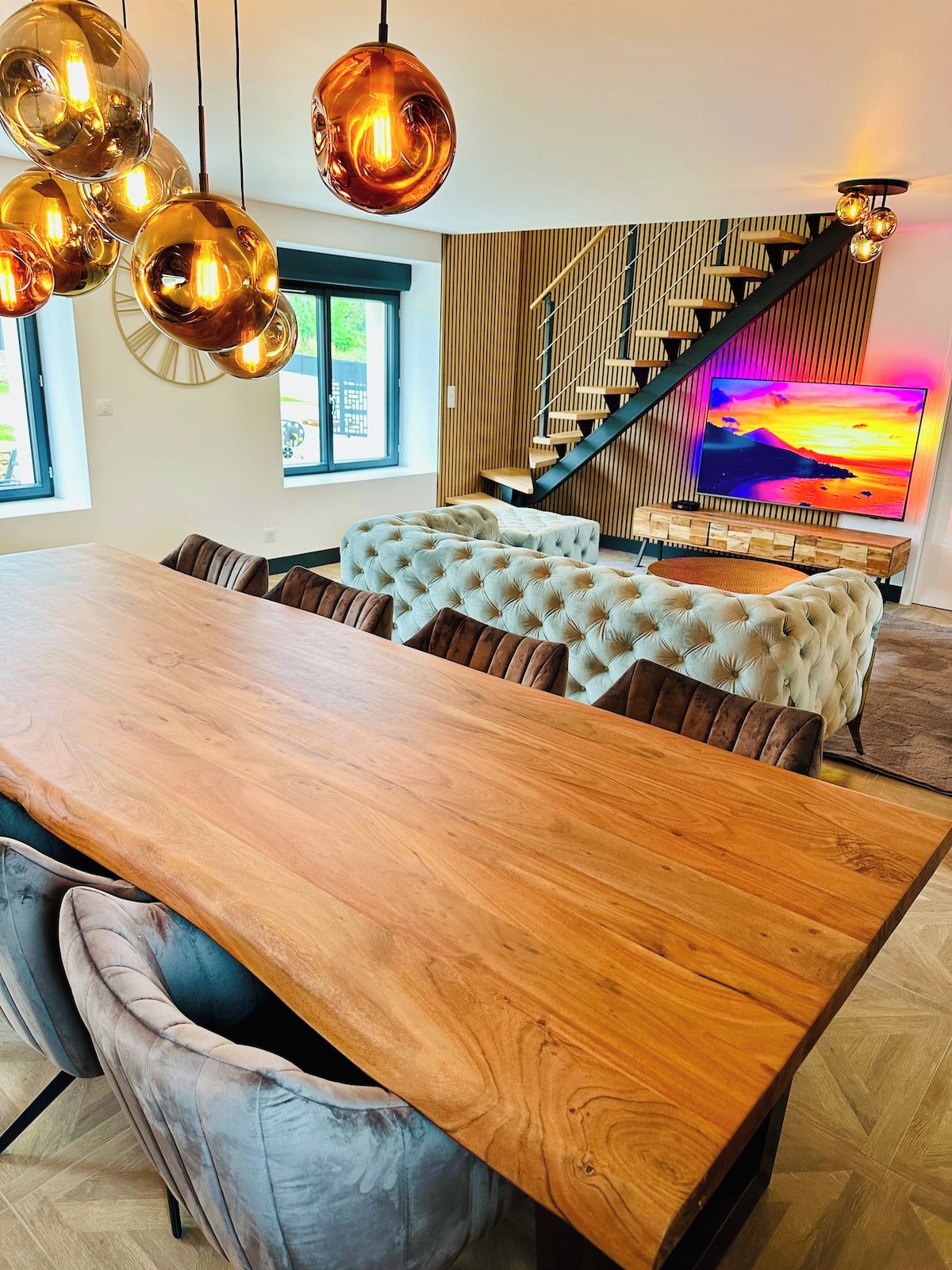 Un espace repas moderne avec une table en bois, des sièges rembourrés et un escalier sur lequel est fixé un téléviseur.