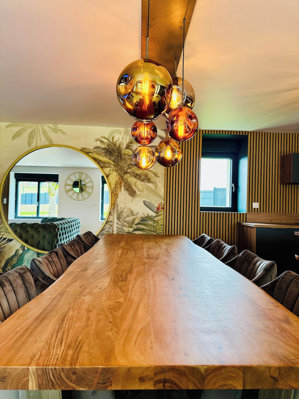 Een eetruimte met een lange houten tafel, gestoffeerde stoelen, een ronde spiegel en een wandlamp.