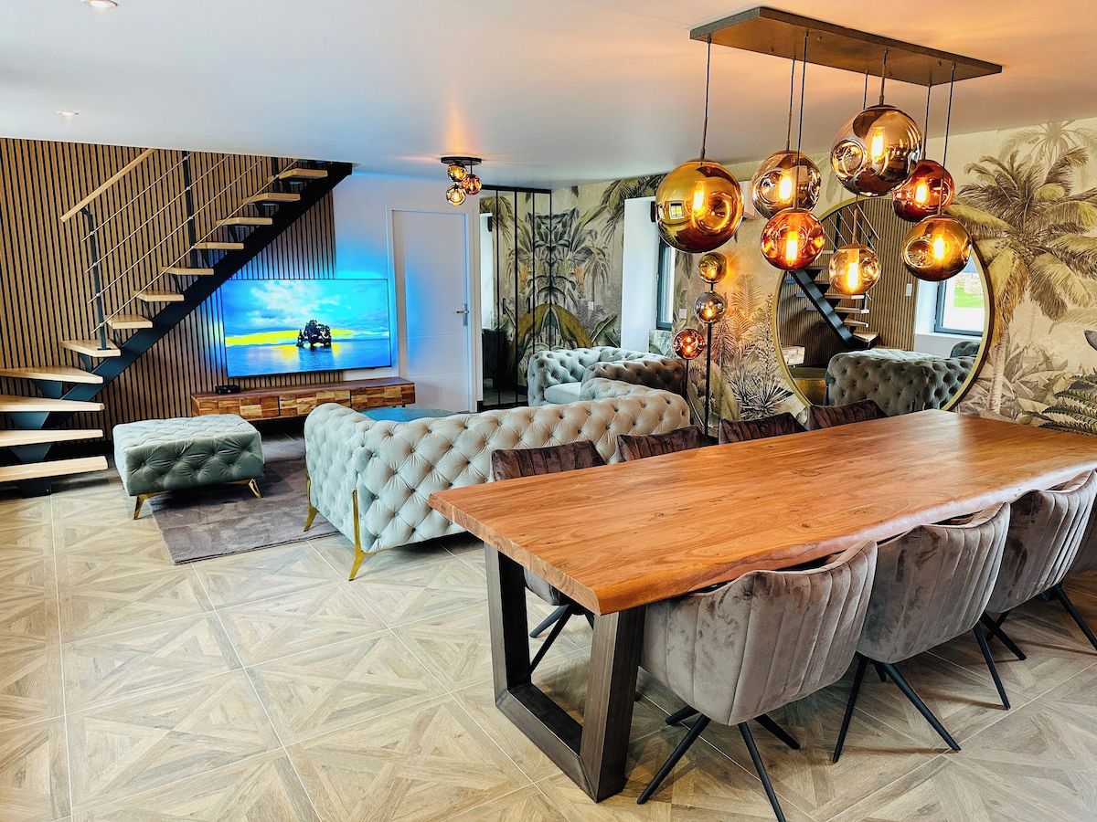 Un salon moderne avec une table à manger en bois, des canapés capitonnés, un escalier et un grand téléviseur fixé au mur.