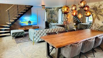 Un salon moderne avec une table à manger en bois, des canapés capitonnés, un escalier et un grand téléviseur fixé au mur.