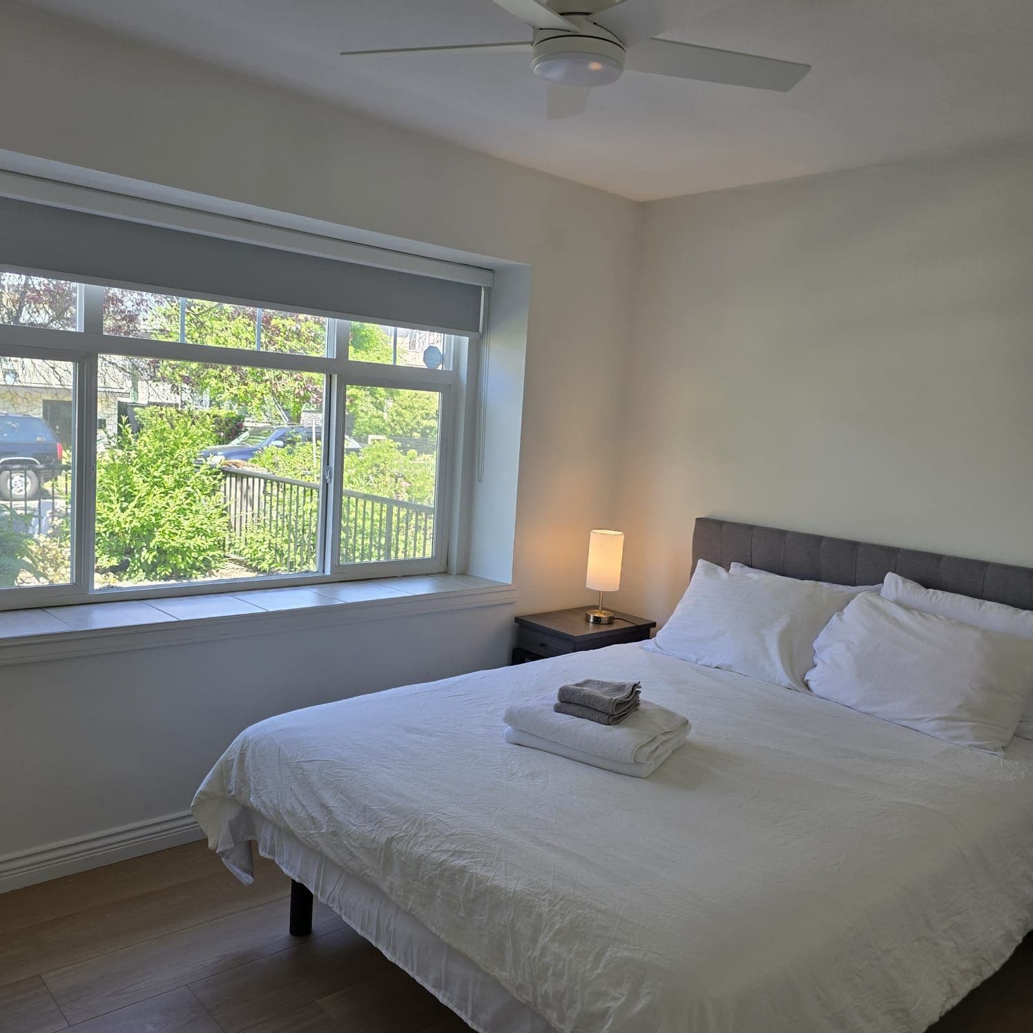 A hotel room with a bed, a nightstand, a lamp, and a window with a view of greenery and a fence.