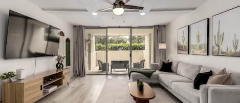 Una sala de estar moderna con un televisor de pantalla plana, un ventilador de techo, un sofá, una mesa de centro y un ventanal corredero que da a un área exterior.