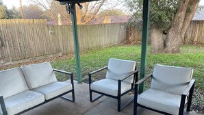 Un patio al aire libre con dos sillas blancas acolchadas y una mesa, rodeado por una valla de madera y árboles.