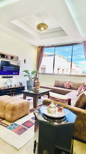 A living room with a flat-screen TV, a sofa, a coffee table with a tray, and a view of a building through large windows. - Super Cozy with Nature Views | Fiber Wi-Fi | 10 min from the Beach (Essaouira)