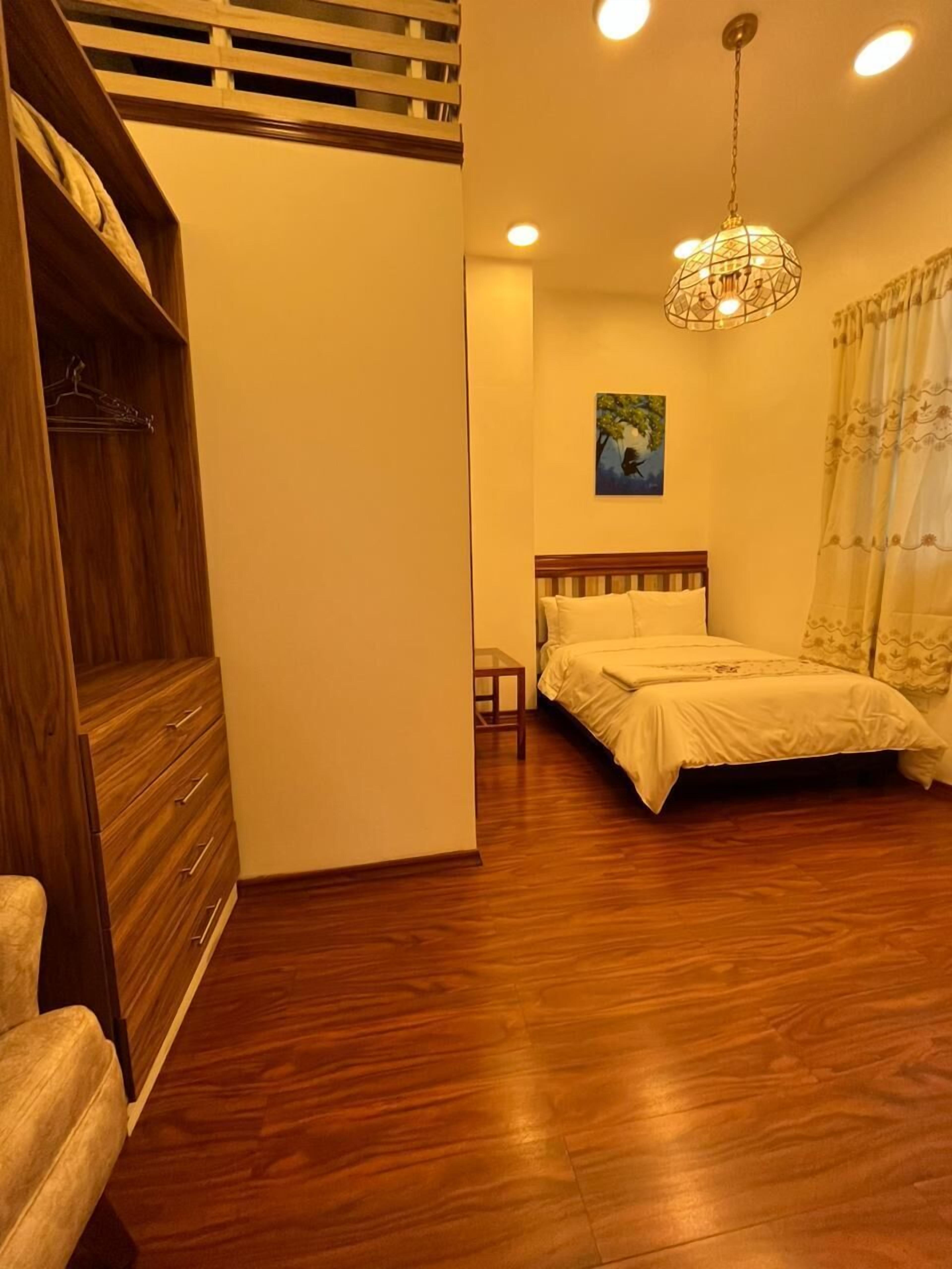 A bedroom with a wooden wardrobe, a bed with white bedding, a hanging light fixture, and a painting on the wall.