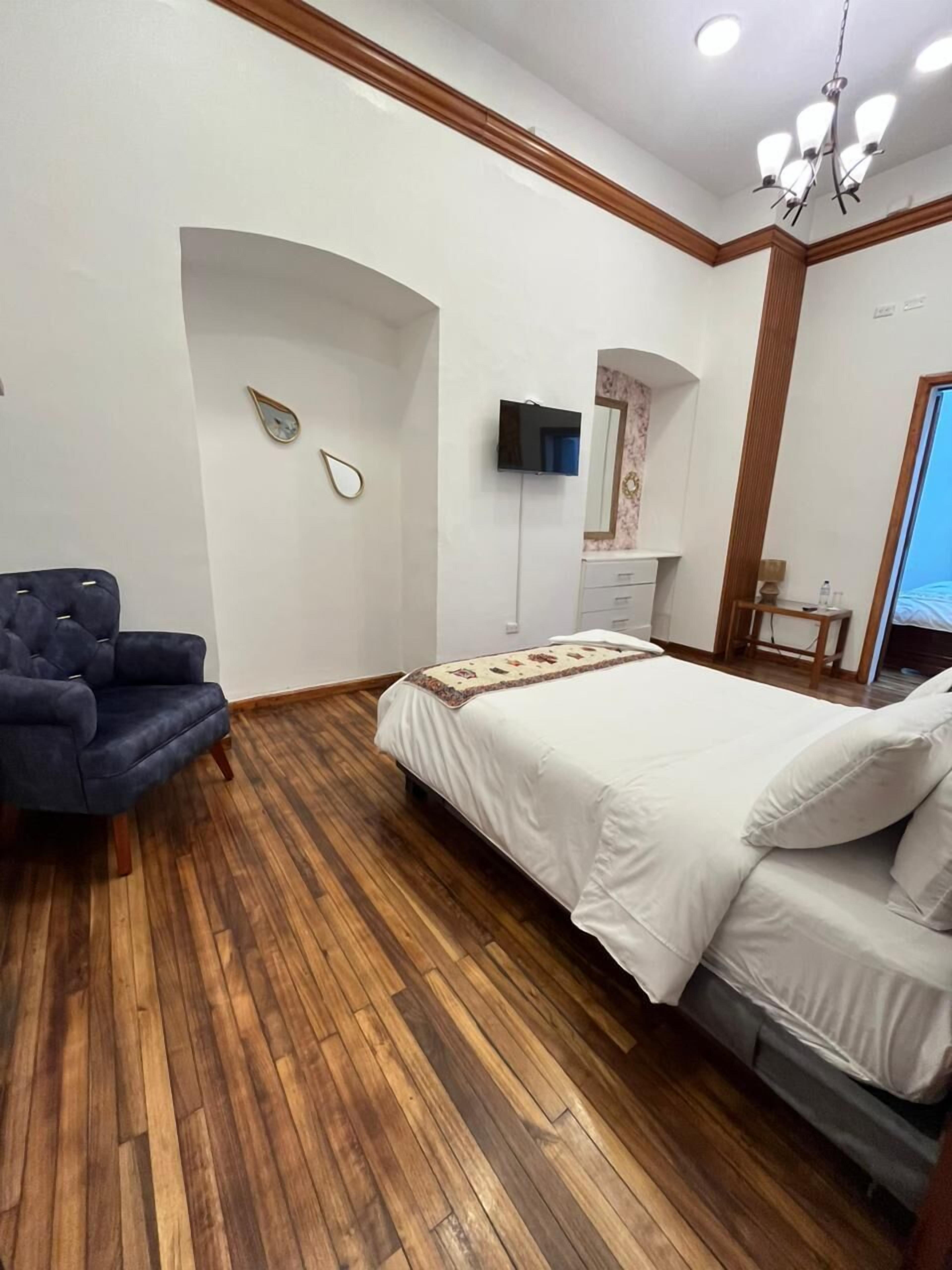 A hotel room with a wooden floor, a bed with white bedding, a blue armchair, a television, and a mirror.