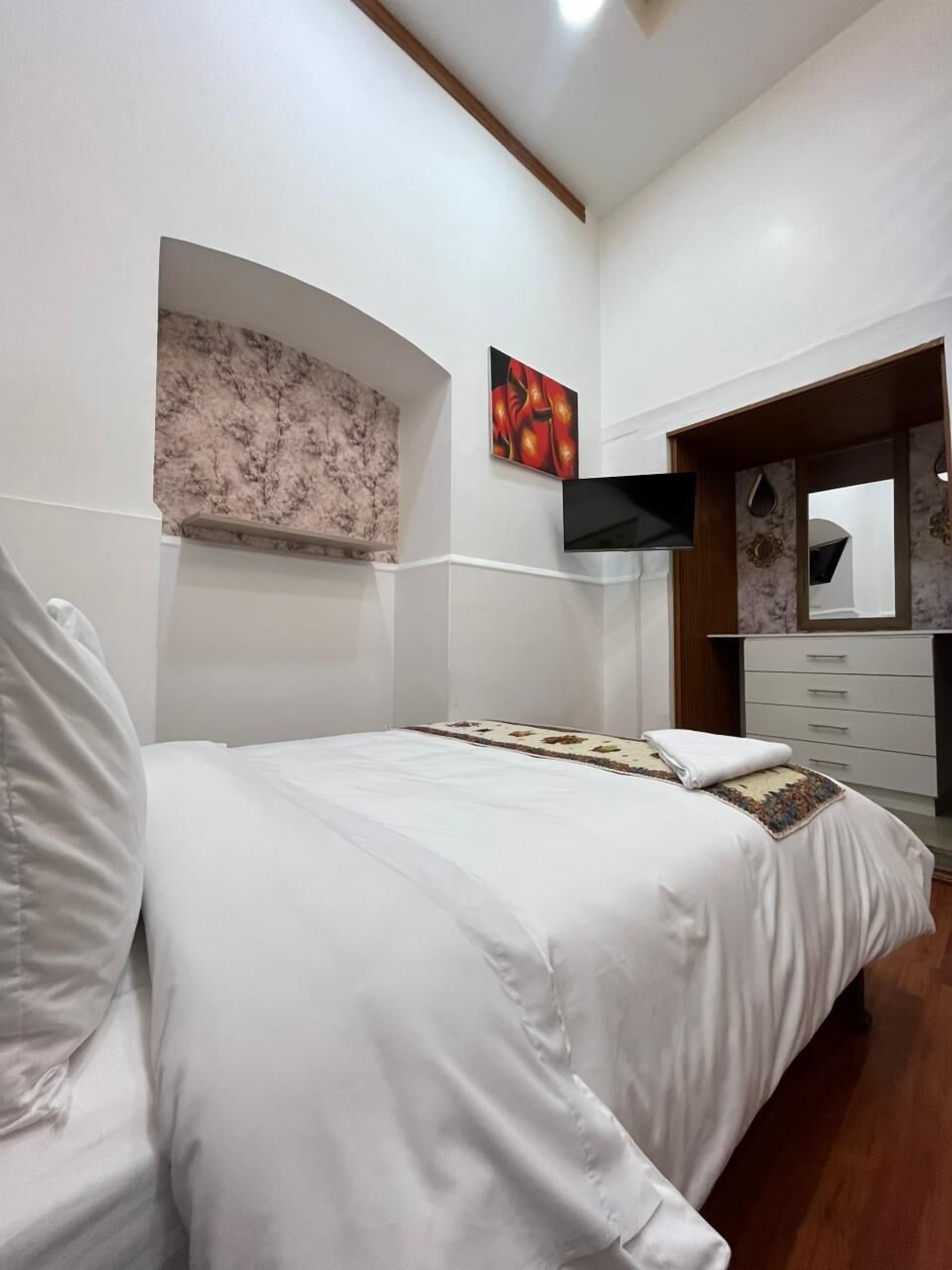 A bedroom with a bed, a dresser, a mirror, and a wall-mounted TV.