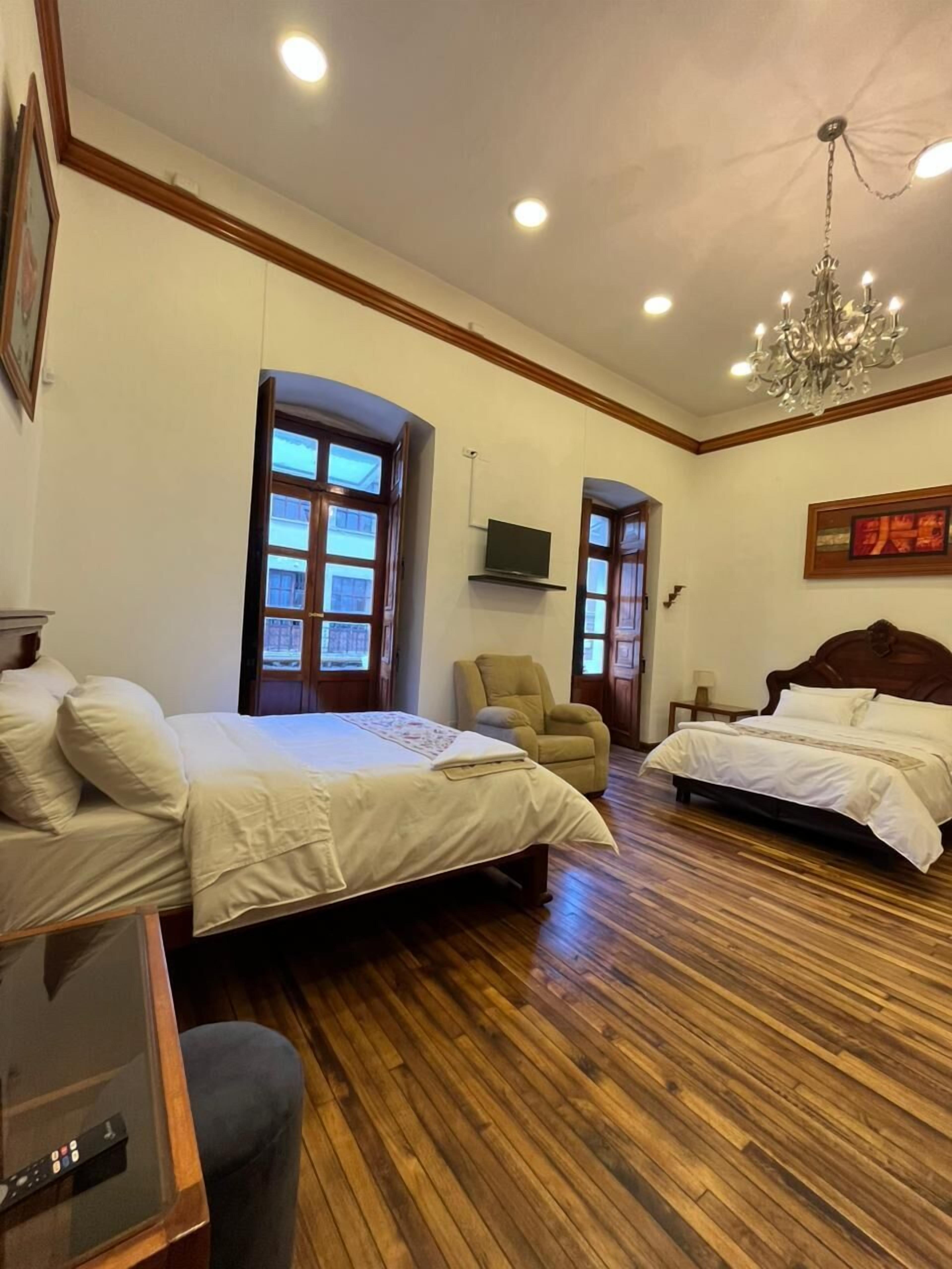 A hotel room with two beds, a TV, a chandelier, and wooden flooring.