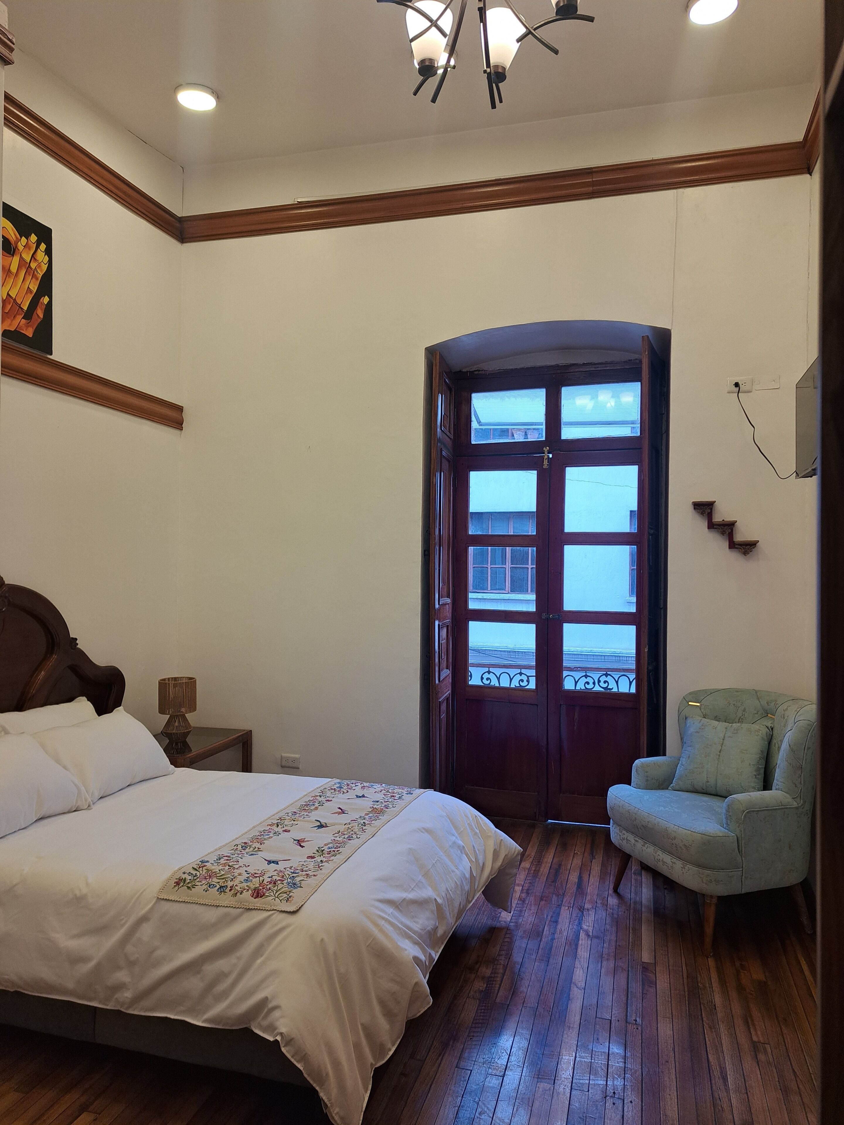 Un dormitorio con cama, una silla, una puerta con ventanas, una lámpara en el techo y un enchufe en la pared.