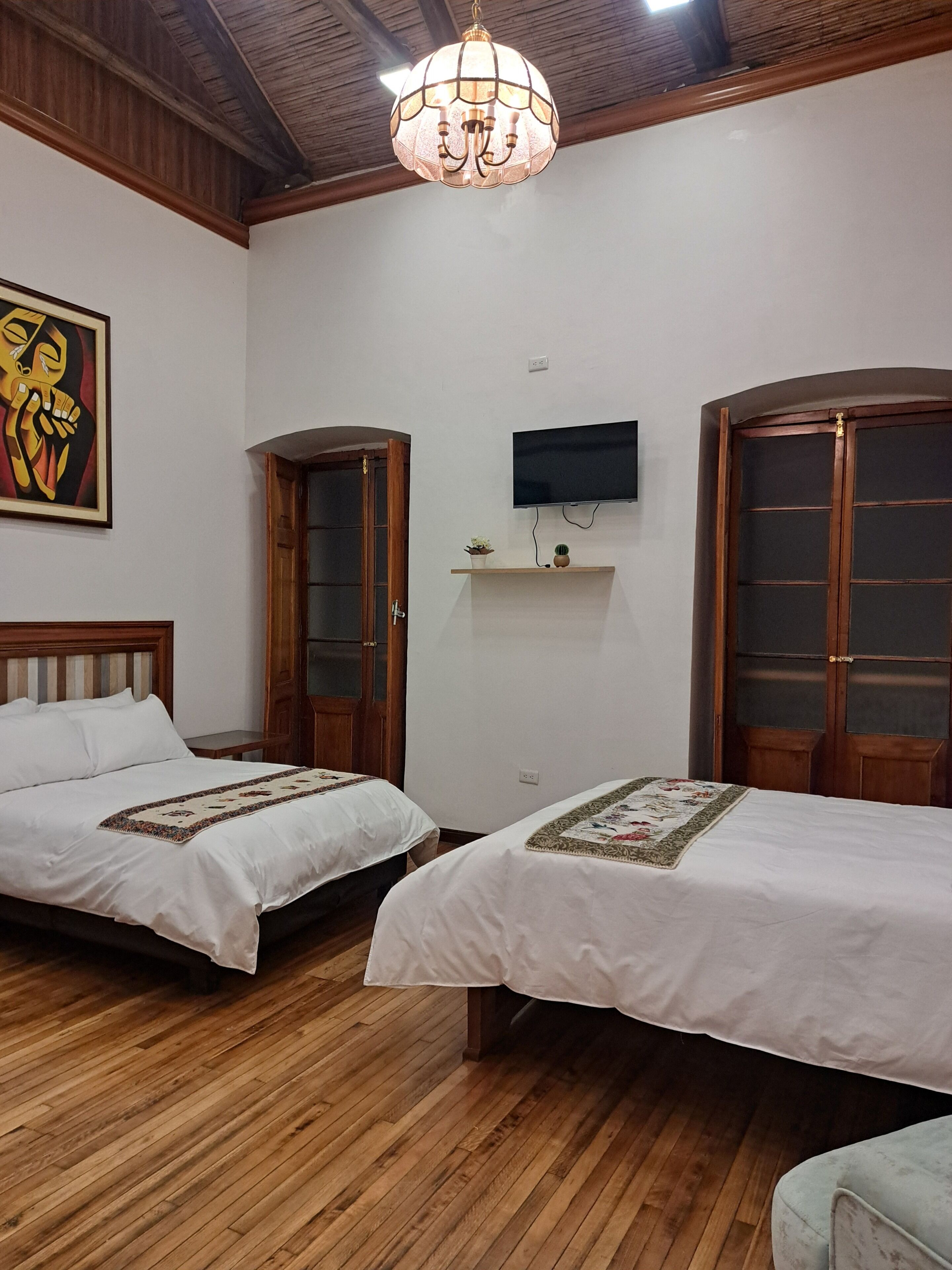 A room with two beds, wooden flooring, a chandelier, a TV, and a painting.