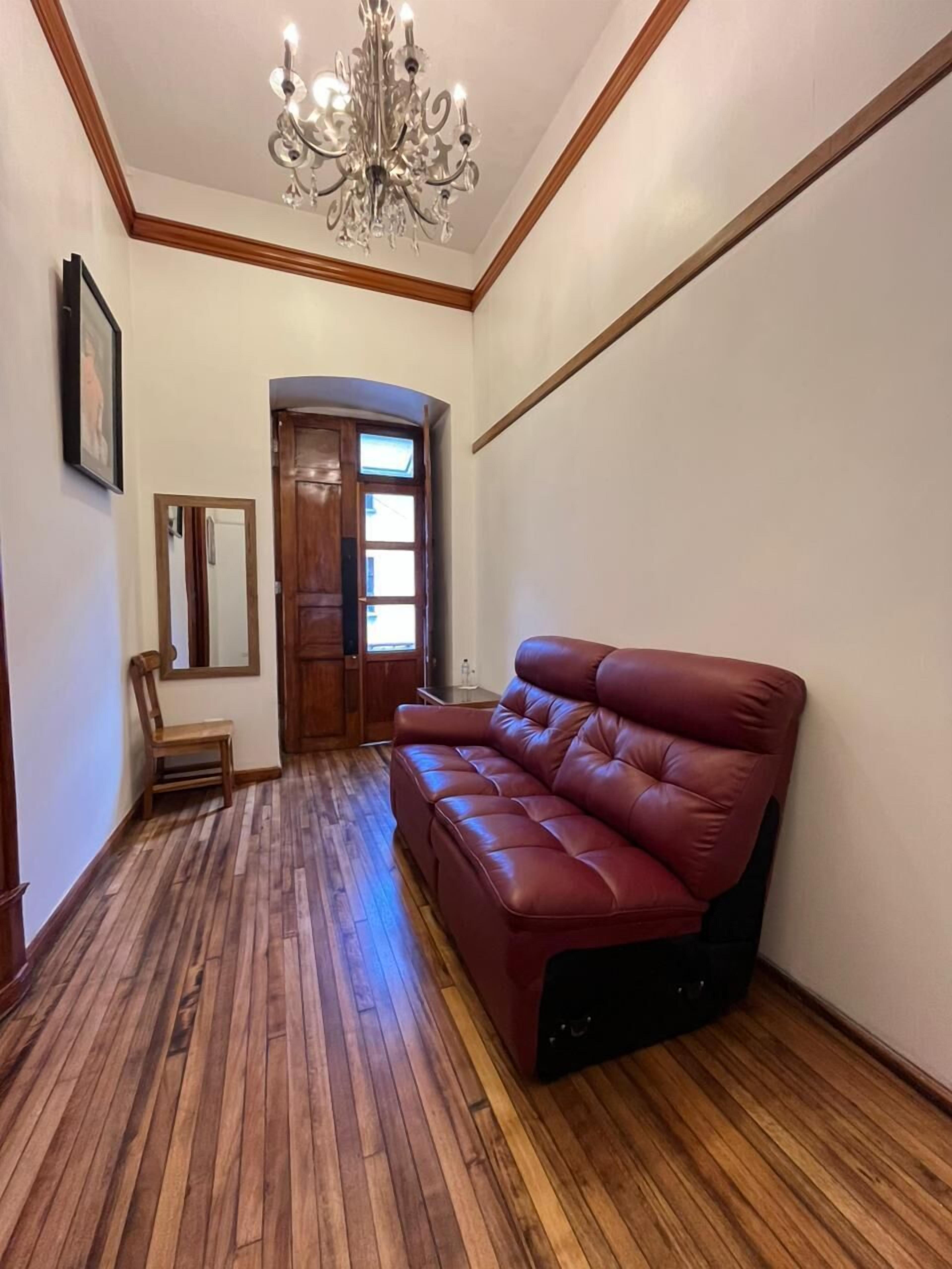 A hallway with a wooden floor, a red leather sofa, a wooden door, a mirror, and a chandelier.