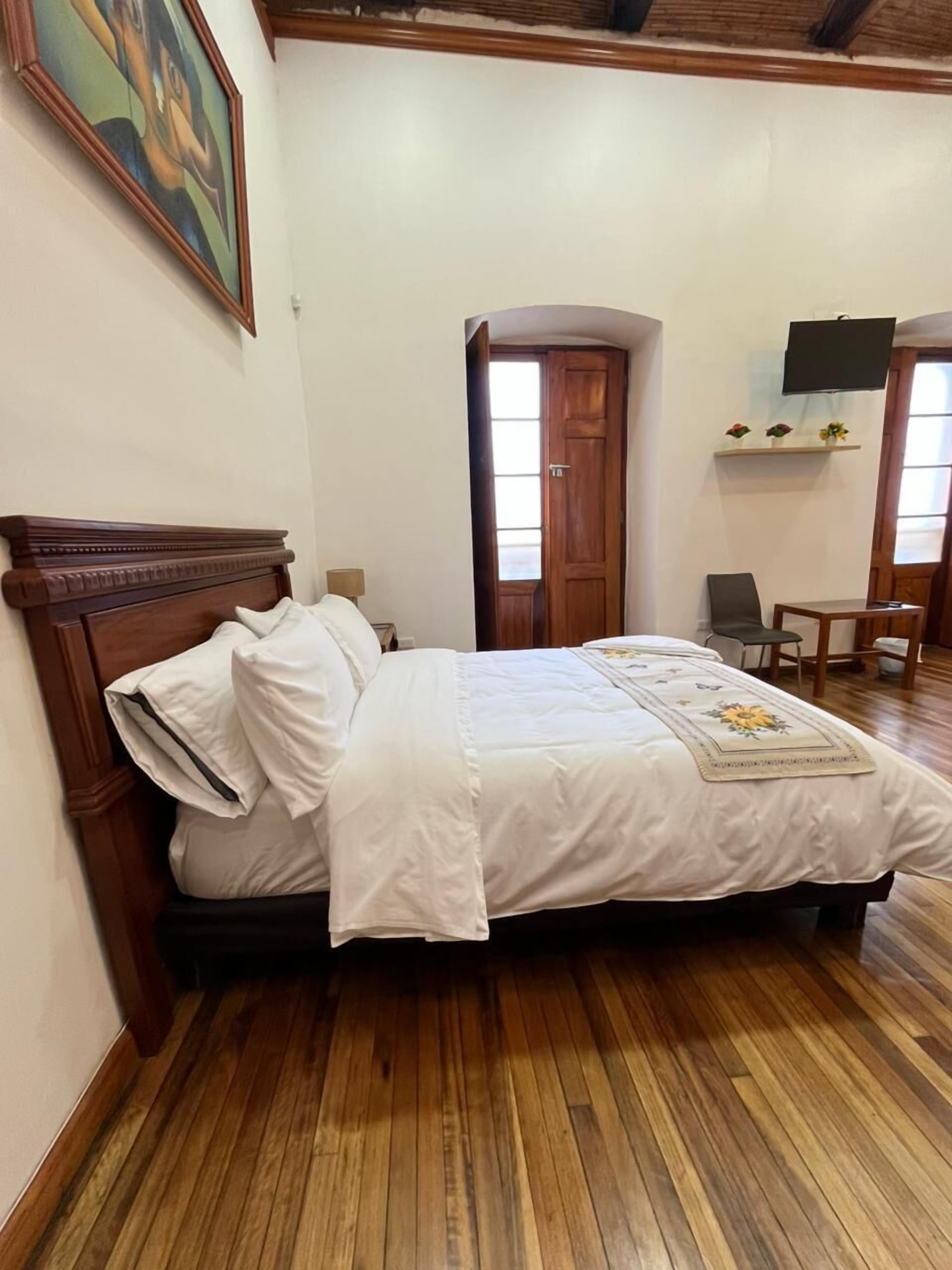 A bedroom with a wooden bed, a nightstand, a chair, a desk, a TV, and a door.