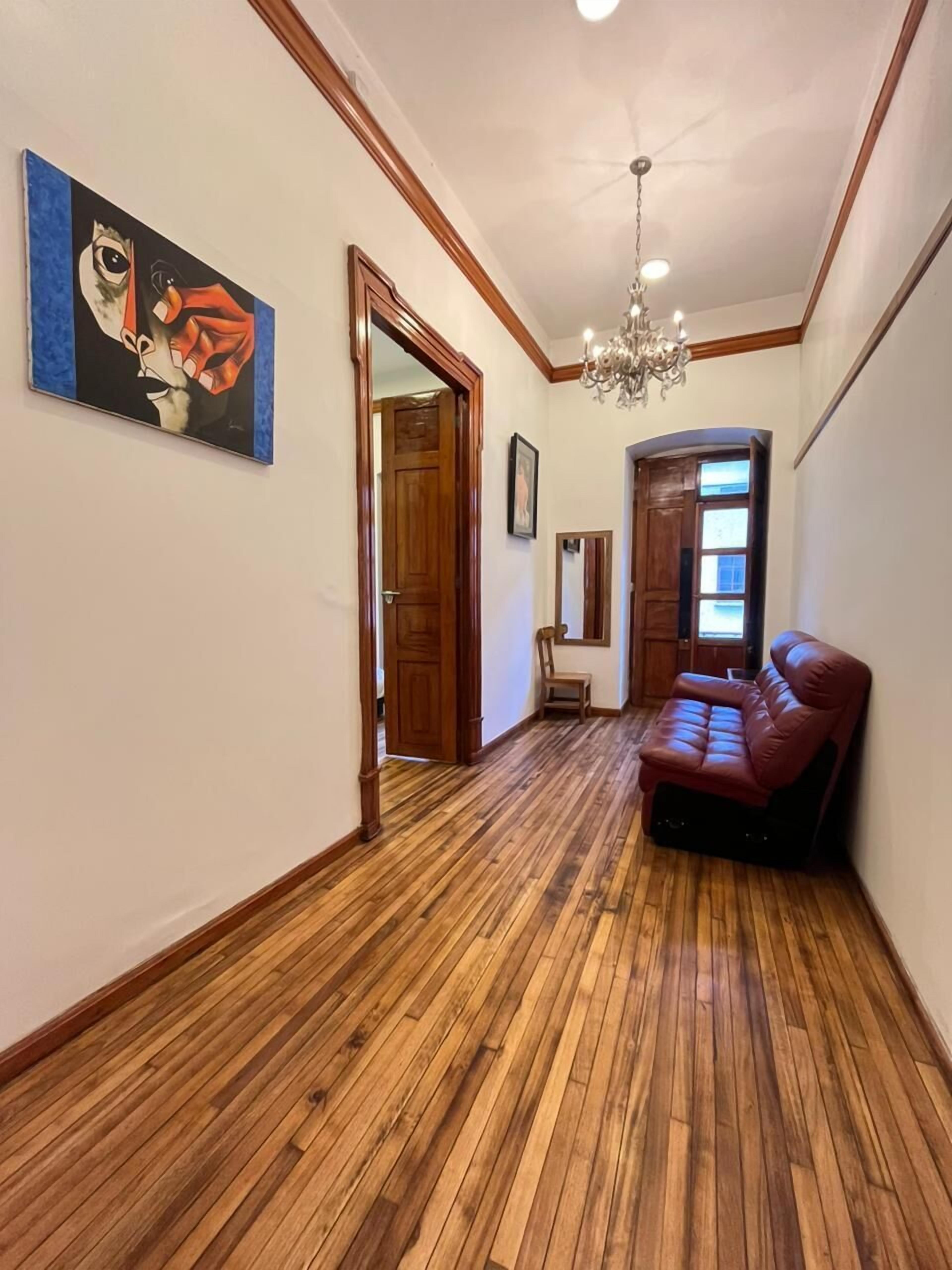 A hallway with wooden flooring, a red leather sofa, a chandelier, and a painting on the wall.