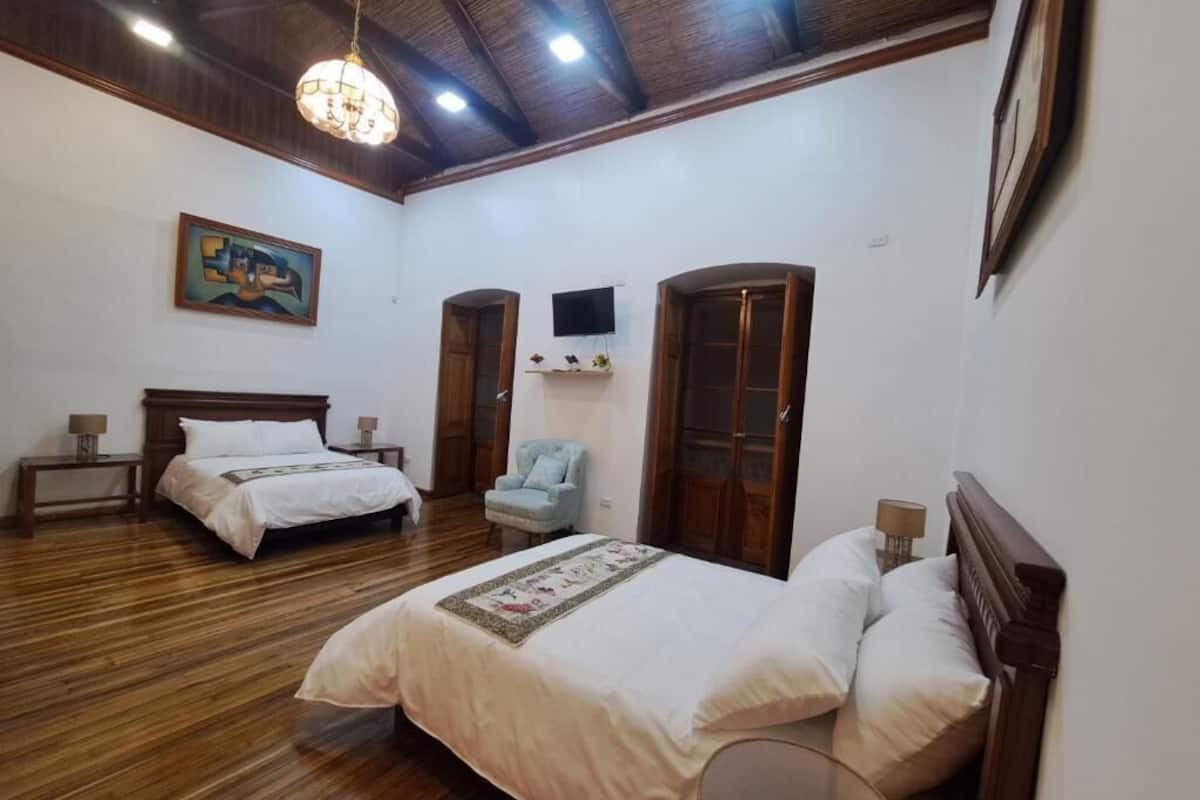 Una habitación con dos camas, piso de madera, un candelabro, un cuadro y un televisor en la pared.