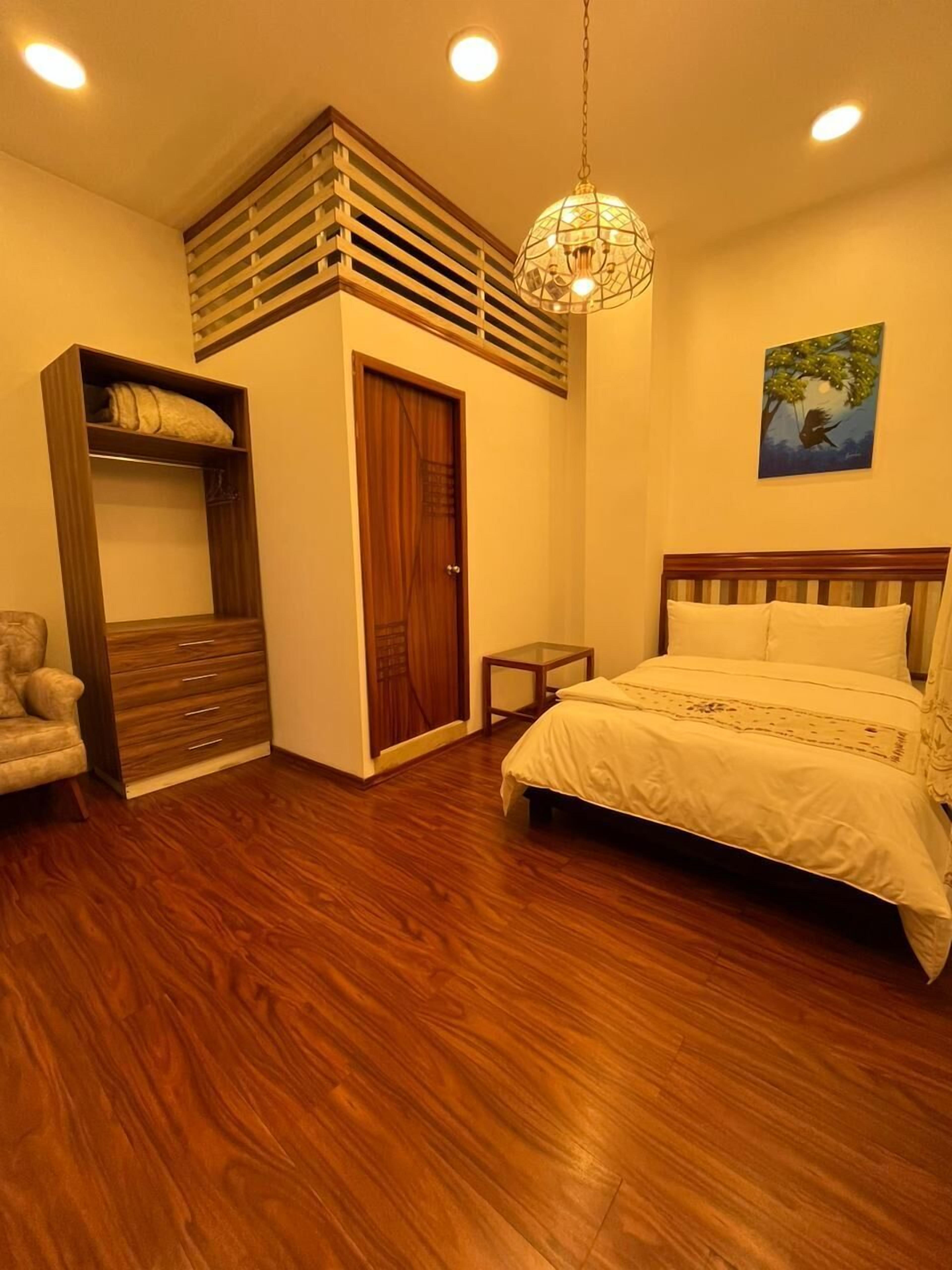 A bedroom with a wooden floor, a bed with white bedding, a wooden wardrobe, a small table, and a hanging light fixture.