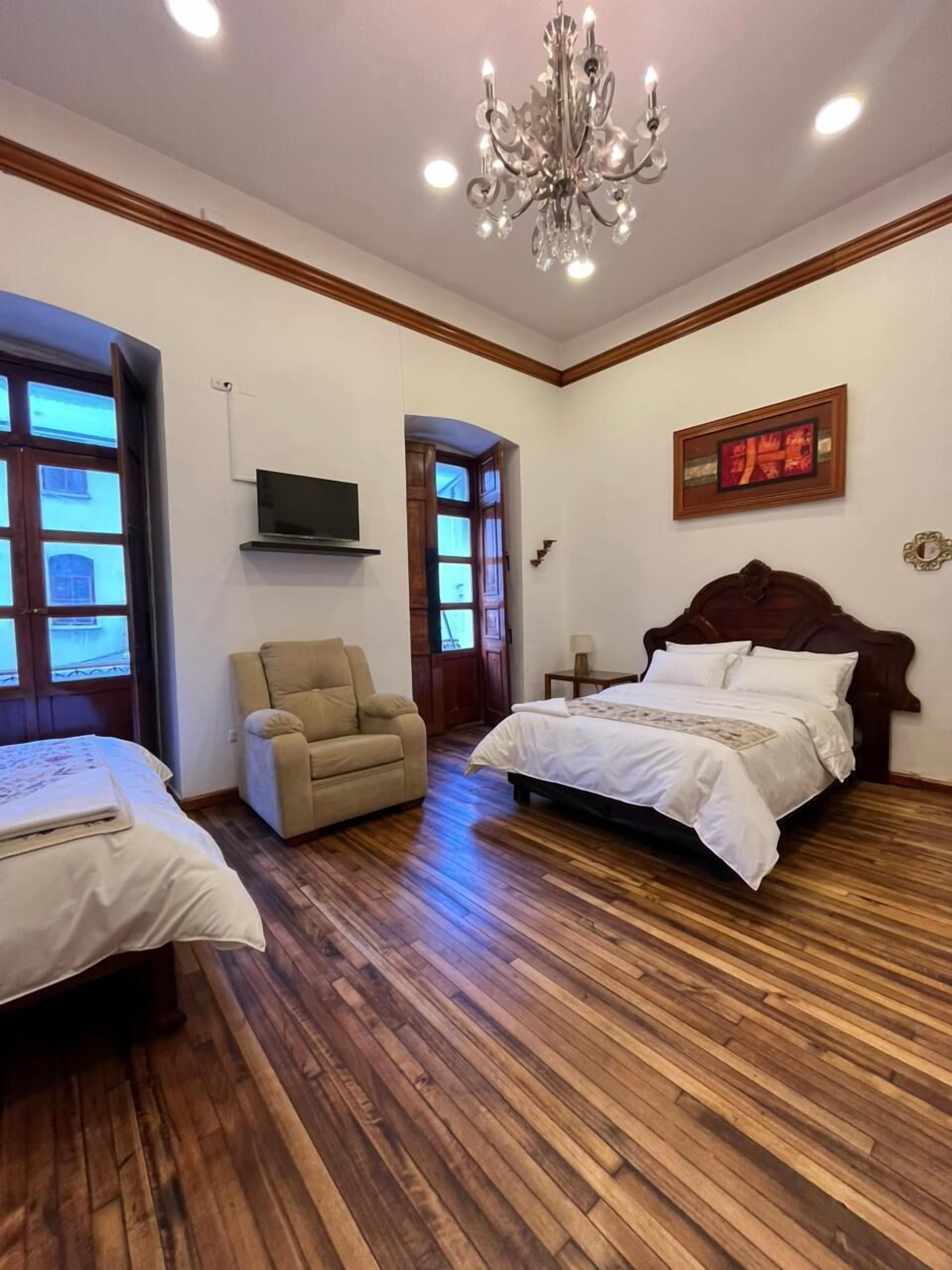 A bedroom with a wooden floor, a bed, a chair, a TV, and a chandelier.