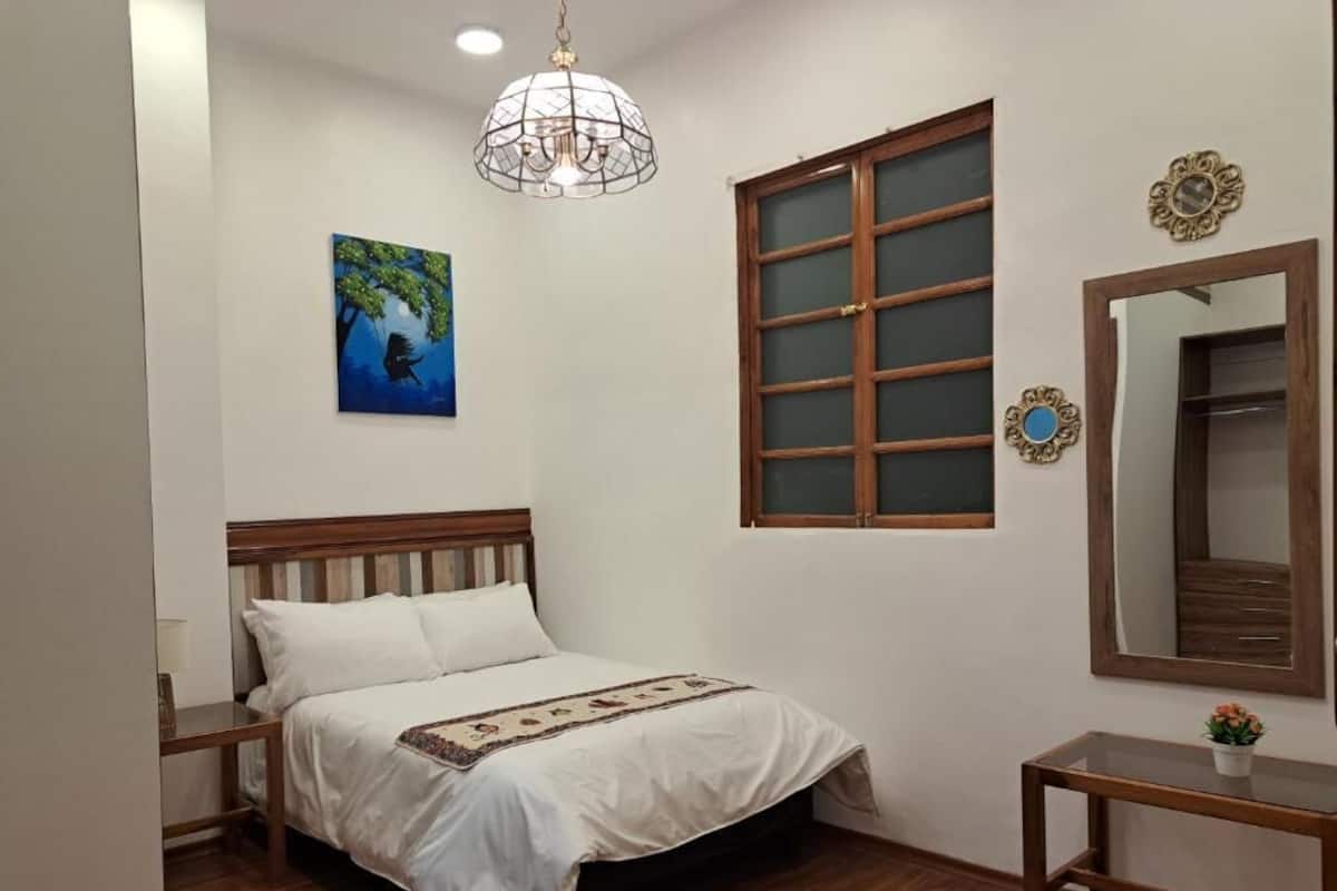 Un dormitorio con cama, mesita de noche, ventana y un cuadro en la pared.
