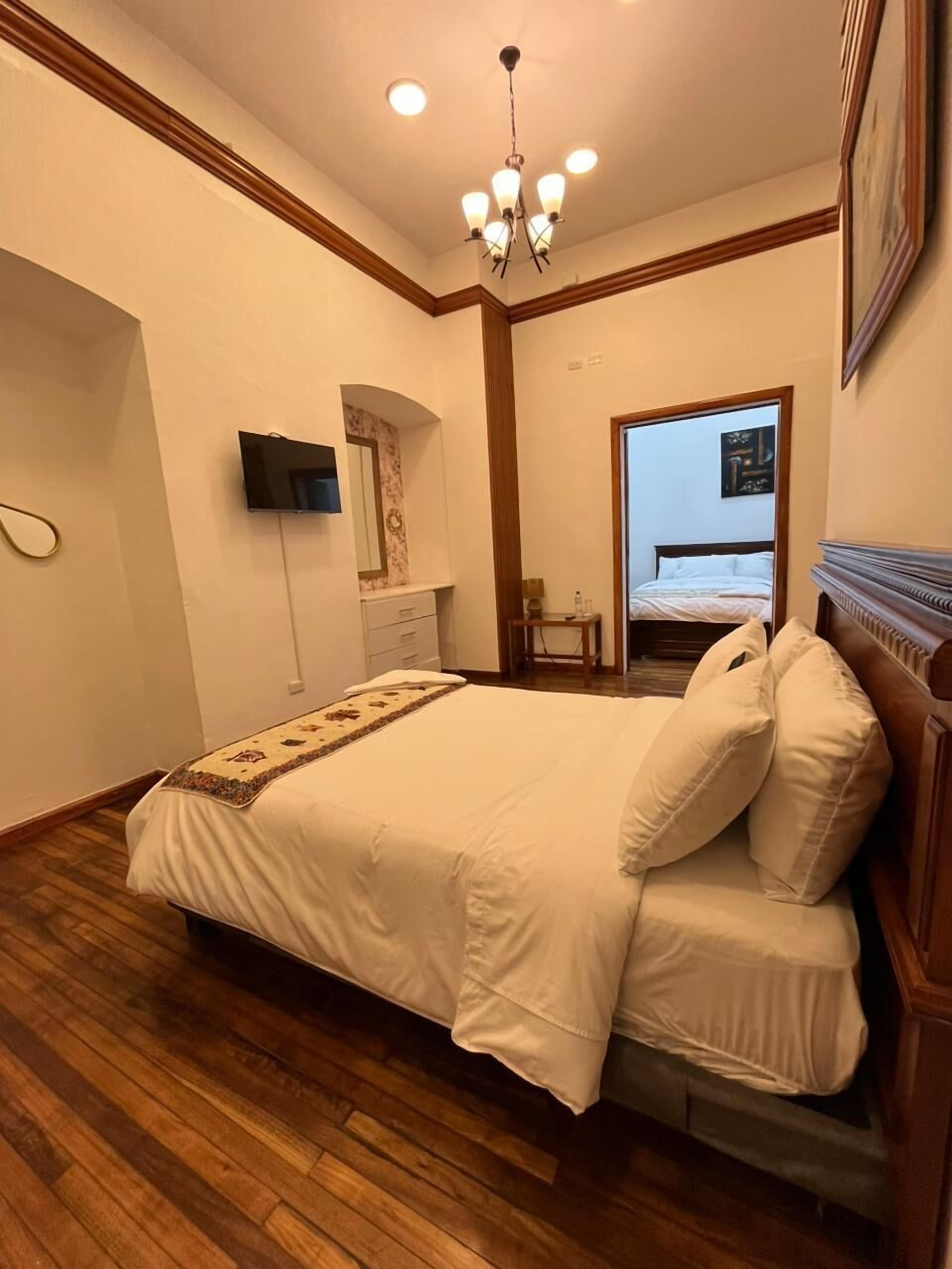 Un dormitorio con suelo de madera, una cama con ropa de cama blanca, un televisor montado en la pared y una araña.