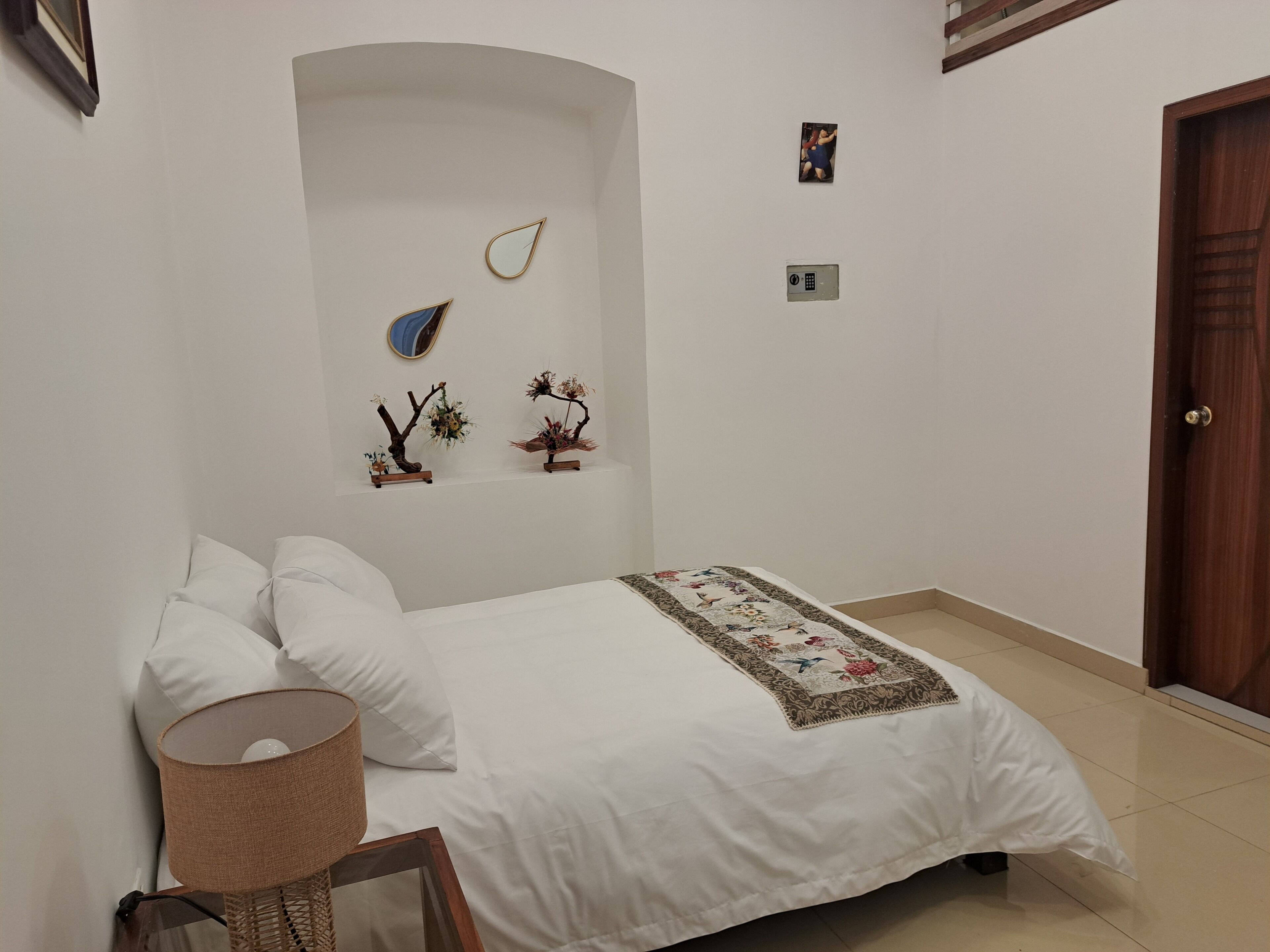 Ein Schlafzimmer mit einem Bett, Kissen, einer Lampe, einem kleinen Regal mit Dekoration und einer Tür.