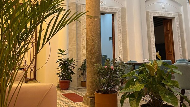 Een hotel lobby met betegelde vloer, potplanten, een zithoek en een trap naar een hoger gelegen verdieping.