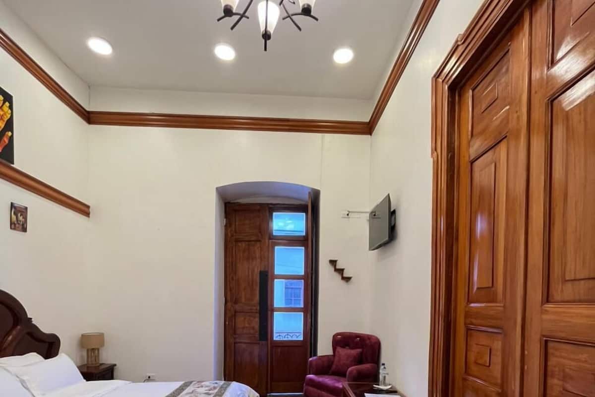 Habitación de hotel con una cama grande, suelo de madera, una araña de cristal, un sillón rojo y un armario de madera.