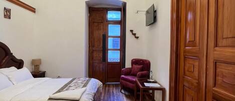 Une chambre d’hôtel avec un grand lit, un parquet en bois, un lustre, un fauteuil rouge et une armoire en bois.