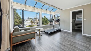 A room with a treadmill, a sofa, a small table, and large windows with a view of houses and trees. - Luxury 5BR Point Grey | Jericho Beach + Downtown (Vancouver)