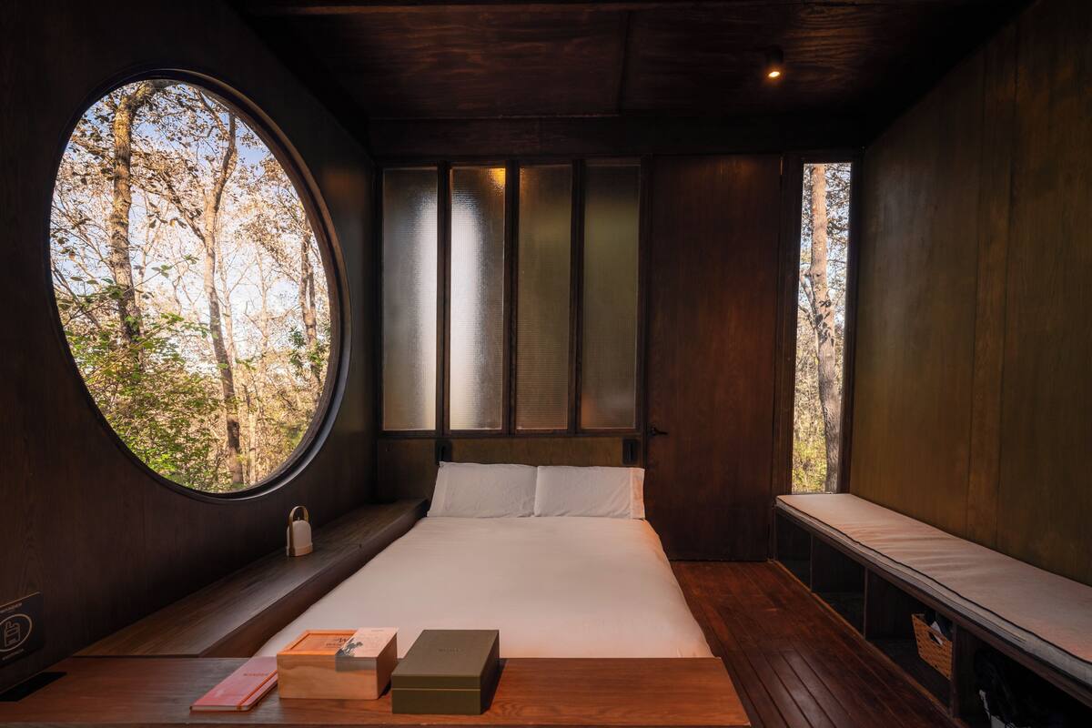 Een slaapkamer met een groot rond raam, een bed met witte lakens, een nachtkastje met boeken en een bankje langs de muur.