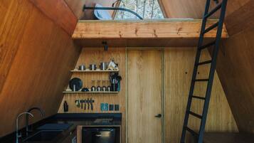 A compact kitchen with wooden cabinets, a sink, and a stove. A ladder is propped against the wall, and a window provides natural light.