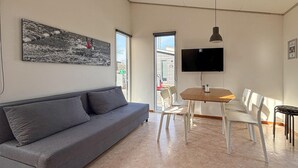 Ett modernt vardagsrum med en grå soffa, ett matbord i trä med vita stolar, en platt-TV som är monterad på väggen och ett stort fönster med utsikt över gatan.
