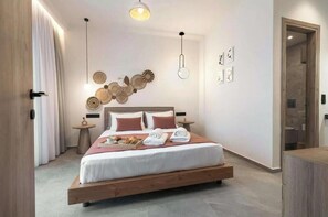 A modern hotel room with a wooden bed, a small bedside table, a wall-mounted light fixture, and a door leading to another room. - Villa Pezoulia Kamilari – Panoramic Luxury Above Crete (Kamilari)