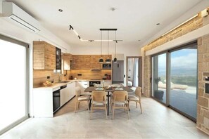 A modern kitchen with a dining area, large windows, and a stone accent wall. - Villa Pezoulia Kamilari – Panoramic Luxury Above Crete (Kamilari)
