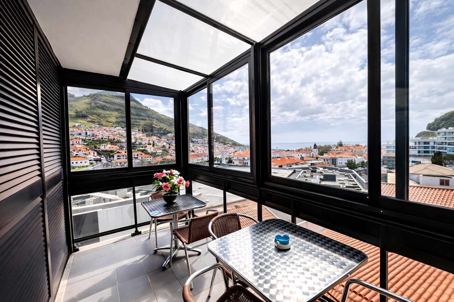 Un balcón con una mesa y sillas, un florero con flores y vistas a un pueblo con edificios de tejados rojos y colinas.
