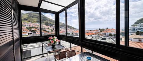 A balcony with a table and chairs, a vase of flowers, and a view of a town with red-roofed buildings and hills.