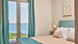 Quarto com cama, mesa de cabeceira com luminária e vista para o mar através de uma porta de vidro.