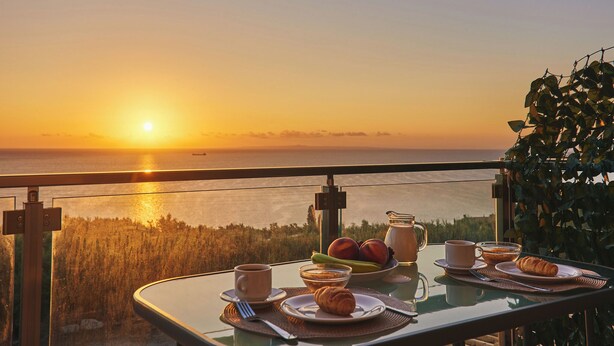 Uma mesa com itens de café da manhã, um copo de suco e um croissant, com vista para o pôr do sol.
