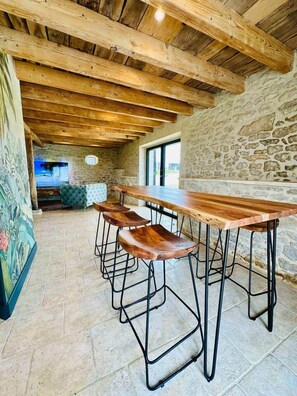 A rustic interior with a long wooden table and bar stools, stone walls, and a stone floor. - Le Belvédère du Quercy - Domaine Gouloumès (Soucirac)