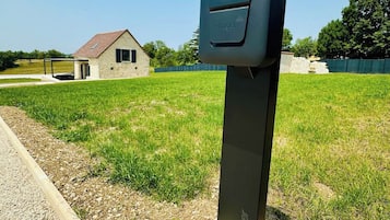 Une structure métallique noire, dotée d’un clavier, qui pourrait être un parcmètre, se trouve dans un champ herbeux, avec un bâtiment en arrière-plan.