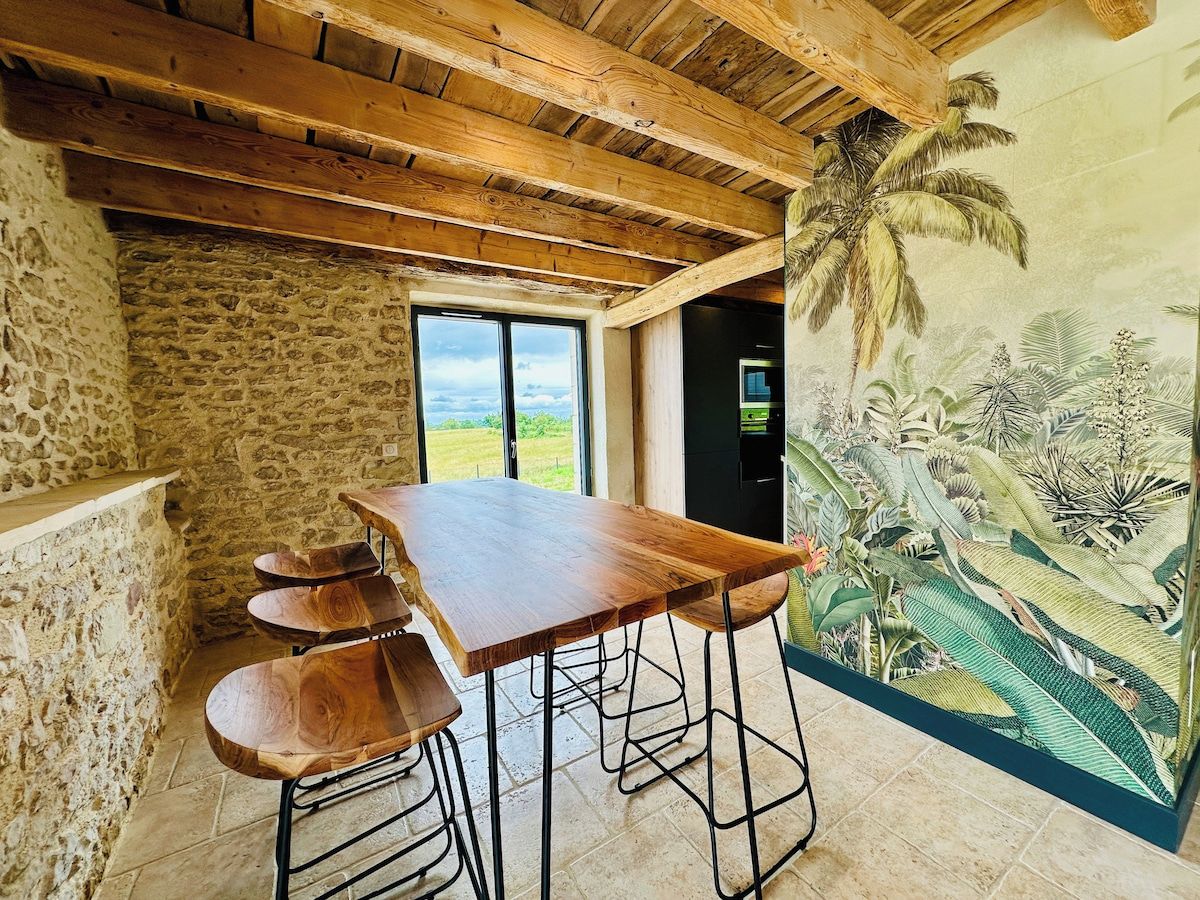 Uma área de refeições rústica com mesa e banquinhos de madeira, paredes de pedra e um mural com folhagem tropical.