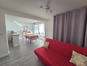 A modern living room with a red sofa, a dining area with a table and chairs, and a kitchen area with a counter and cabinets. - CAP D'AGDE - Plage du Môle // Réf 0208 (CAP D'AGDE)